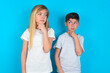 © Jihan - Dreamy two kids boy and girl standing over blue studio background with pleasant expression, looks sideways, keeps hand under chin, thinks about something pleasant.