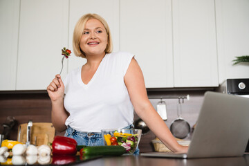 Low angle view plus size plump woman culinary vlogger blogger eating vegetable salad, preparing healthy vegan vegetarian food using laptop for blogging online at home kitchen