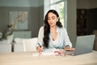© Eva March - Smiling successful female using laptop computer indoors at home office, making notes on papers, remote work and small business freelancer, education and student.
