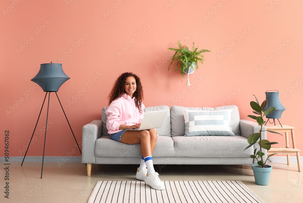 Young woman working with laptop on comfortable sofa near pink wall