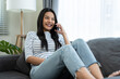 © Kawee - Asian beautiful woman sit on sofa and talk on mobile phone in house. Happy attractive young girl spend leisure time at home, feel relax and enjoy communicate and discuss on smartphone in living room.