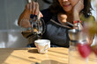 © bongkarn - Cropped, aged woman poring hot coffee into a ceramic cup on the table.