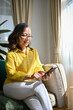 © bongkarn - Happy asian retired woman sitting on comfy couch in living room, using smartphone