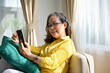 © bongkarn - Cheerful asian aged-woman resting on the sofa and using modern mobile phone.