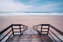 Wooden Stairs Heading Down To The Oceanbeach As Sea Foam And Waves Roll In. Portugal.