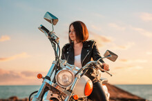 Defocused Portrait Of A Beautiful Adult Woman Driving A Motorcycle. Sunset Sky And Ocean In The Background. The Concept Of The Motorcyclist's Day And Motorcycle Trips
