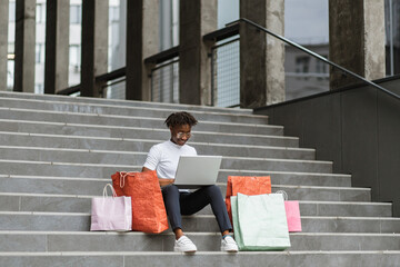 Wall Mural - City shopping concept. Outdoor urban lifestyle portrait of diverse young African American woman, sitting on the steps of the mall with a laptop and colorful shopping bags while shopping online.