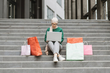 Wall Mural - Outdoor full length fashion portrait of young muslim lady in hijab and stylish clothes sitting on the steps of a large shopping mall and ordering clothes online colored shopping bags lie next to her.