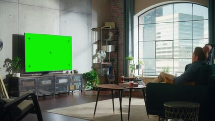 Aufkleber - Young Handsome Man Sitting Down on a Sofa and Watching TV with Horizontal Green Screen Mock Up. It's Day Time in Room at Home Living Room in Loft Apartment.