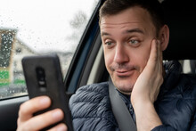 Millennial Man Shocked When See Smartphone While Sitting In Car. Surprised Businessman Look At Phone Screen In Taxi. Unshaven Man In Blue Jacket Holds Phone In His Hands And Looks In Surprise In Car.