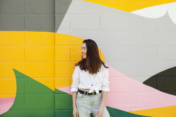 Young beautiful millennial woman standing confidently by bright colorful green, yellow, pink wall, smiling and laughing - candid lifestyle ordinary moments