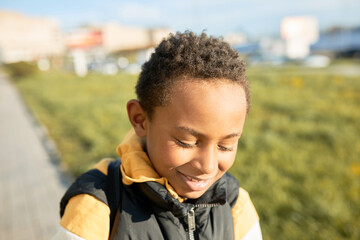 Wall Mural - Horizontal outdoor portrait of shy african kid in hoodie and puffer vest looking down posing over cityscape with green lawn and sidewalk, walking in sunny evening. Happy carefree childhood