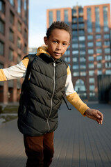Wall Mural - Cool stylish african american kid in hoodie and puffer vest jumping over multi-storey buildings in his residential area, waiting his friend to go for walk after returning home from school