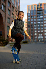 Active black cute boy of 8-years-old in stylish outfit doing tricks on blue penny board over residential area buildings. Urban lifestyle. Happy childhood, big city life and outdoor activities