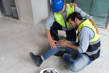 Young caucasian man in safety vest gripped his knee and screams in pain. Due to injuries at the construction site. His asian friend comes and help him to stand.