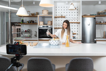 Young vlogger woman vlogging about cooking at her apartment domestic kitchen cook and making video vlog with smartphone as online content creator for internet. Female influencer making a meal.