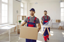 Workers From Moving Company Or Truck Delivery Service Removing Furniture And Other Things From House Or Apartment. Two Happy Young Men In Workwear Uniforms Together Carrying Boxes And Chairs