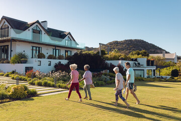 Wall Mural - Multiracial male and female seniors walking on grassy land towards nursing home against clear sky