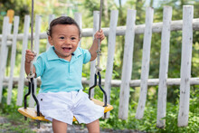 Beautiful Smiling One Year Old Baby Riding On A Swing. Cute Little Boy Wears White And Blue While Playing In A Park Full Of Nature.