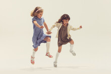 Portrait Of Two Cheerful Happy Girls, Children In Beautiful Dresses Playing Together, Jumping Isolated Over Grey Studio Background
