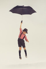 Wall Mural - Portrait of little boy, child in checkered shirt and shorts playing, jumping with umbrella isolated over grey studio background