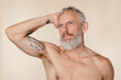 © InsideCreativeHouse - Sexy caucasian strong middle-aged mature man with grey hair and beard shirtless with bare shoulders with tattoos isolated in beige background. Male beauty concept