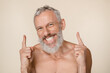 © InsideCreativeHouse - Handsome caucasian mature middle-aged man with grey hair and beard shirtless looking at camera pointing showing copy space free space isolated in beige background