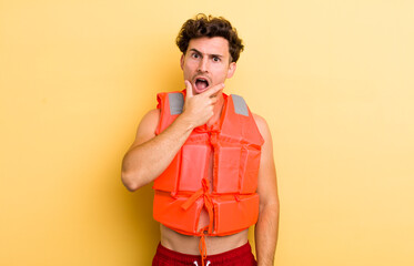 Wall Mural - young handsome guy with mouth and eyes wide open and hand on chin. life jacket and boat concept