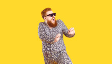 Crazy Plus Size Man In Funny Pajamas Dancing And Having Fun In Fashion Studio. Happy Bearded Fat Guy Wearing Cool Sunglasses And Comfy Leopard PJs Singing And Dancing Isolated On Yellow Background