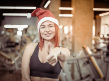 Attractive Red-haired Fitness Woman In Santa Hat Showing Thumb Up In The Modern Gym. Christmas, New Year, Fitness Concept