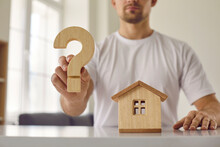 Confusion Young Man Who Is Hesitant About Buying His Home Shows Wooden Question Mark. Close Up Of Question Mark And Wooden House In Hands Of Man Sitting At Table. Concept Of Housing Development.