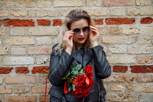 Portrait Of Young Beautiful Woman In Black Dress, Leather Jacket Holding Bouquet Of Red Roses, Standing At Brick Wall.