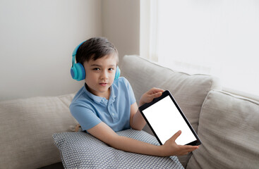 Wall Mural - Kid wearing headphones holding tablet mockup, School boy using digital pad doing homework,Child sitting on sofa playing game online on internet with friend,Children with technology and Social media