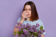 © sementsova321 - Unhappy dark haired woman suffers from runny nose uses nasal spray, has allergic rhinitis reaction on lilac flowers, posing against purple background.