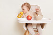 Image of thirsty toddler baby girl dresses in beige jumper sitting in high chair and drinking water from yellow bottle, looking aside, studying area around her, posing isolated over white background