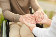© Mediaphotos - Close-up of unrecognizable social care worker and disabled patient in wheelchair holding hands