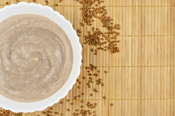Wall Mural - Buckwheat porridge for a baby from ground cereals in a white bowl on a brown bamboo background. Space for text. Baby's first complementary food, baby nutrition.