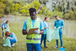 © Dragana Gordic - African volunteer man with clipboard in park. Africa volunteering, charity, people and ecology concept. Behind him are volunteers cleaning the public park...