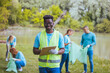 © Dragana Gordic - Medium shot of young African volunteer with clipboard. He is with his team outside in the park, with bags, gloves and buckets, cleaning and collecting garbage