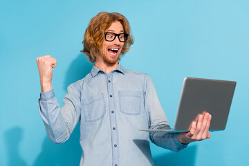 Poster - Photo of funny lucky young guy dressed jeans shirt eyewear rising fist chatting modern device isolated blue color background