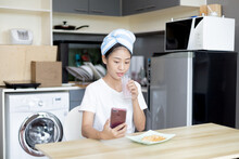 Asian Woman Eats Breakfast With Crackers (Healthy Whole Grain) And Juice, Use Your Phone Or Mobile Phone To Search For Morning News, Small Room In Condominium Background, Wake-up Activities.