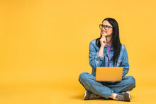 Business Concept. Portrait Of Happy Young Woman In Casual Sitting On Floor In Lotus Pose And Holding Laptop Isolated Over Yellow Background.