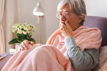 Suffering elderly adult caucasian woman with cough and fever as seasonal flu or pollen allergy sitting on sofa at home