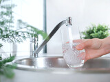 Fototapeta Desenie - A stream of clean water drink flows into the glass. Woman holding a glass of water under running water from the tap in the kitchen.