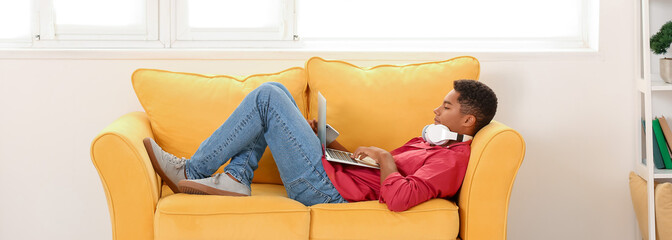 Wall Mural - Teenage African-American boy with laptop and headphones lying on sofa