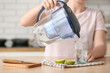 Woman pouring purified water into glass from filter jug at table in kitchen