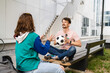 © Miljan Živković - Couple woman and young caucasian man modern happy adult male and female boyfriend and girlfriend smile sitting in front of stadium holding soccer ball outdoor in day football game concept copy space