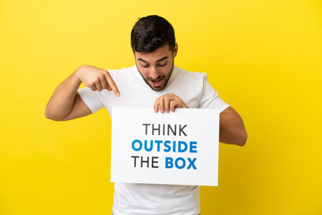 Wall Mural - Young handsome caucasian man isolated on yellow background holding a placard with text Think Outside The Box and  pointing it