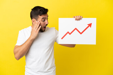 Wall Mural - Young handsome caucasian man isolated on yellow background holding a sign with a growing statistics arrow symbol with surprised expression