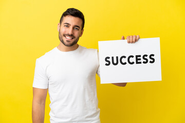 Wall Mural - Young handsome caucasian man isolated on yellow background holding a placard with text SUCCESS with happy expression